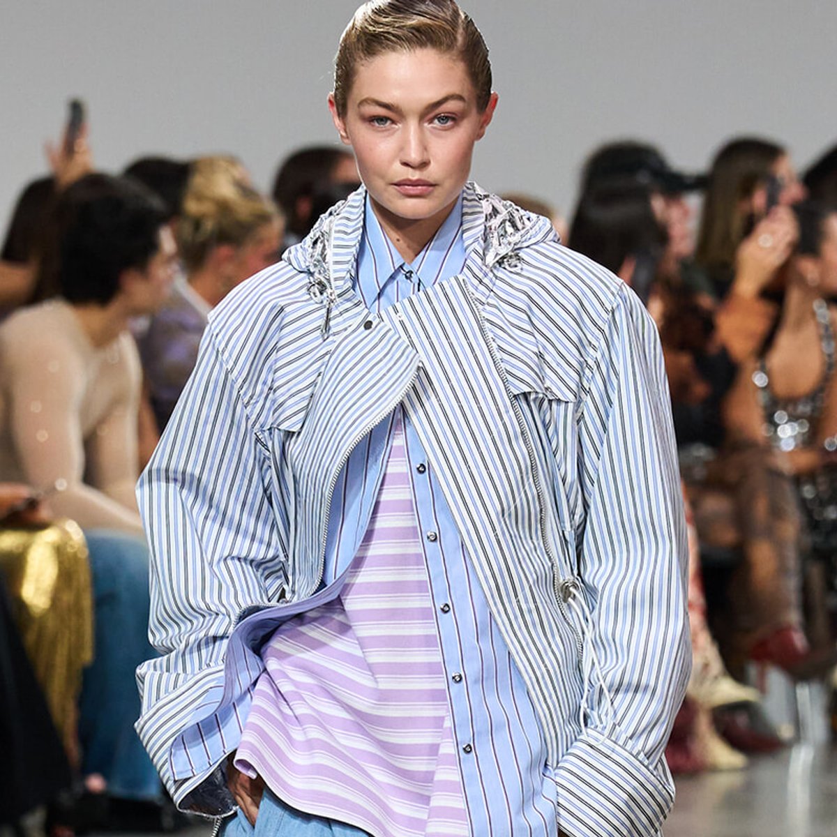 Model walking a fashion show wearing a light blue and white striped oversized jacket over a lavender striped shirt, audience blurred in the background.
