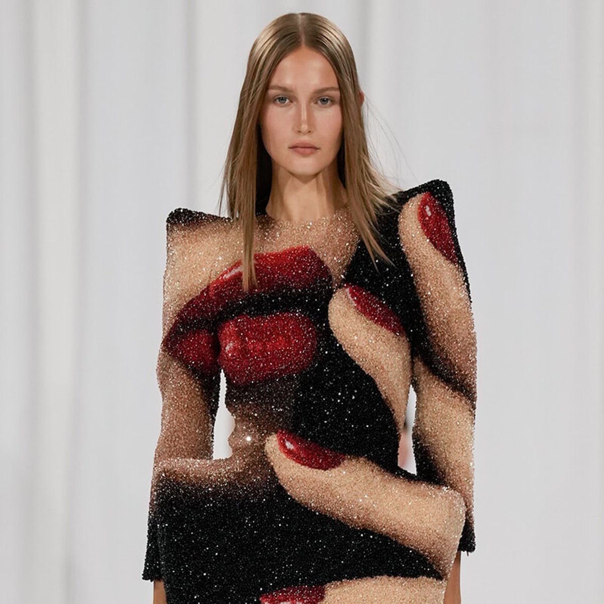 Model wearing a sheer, black and red beaded dress with an abstract pattern on a white backdrop.