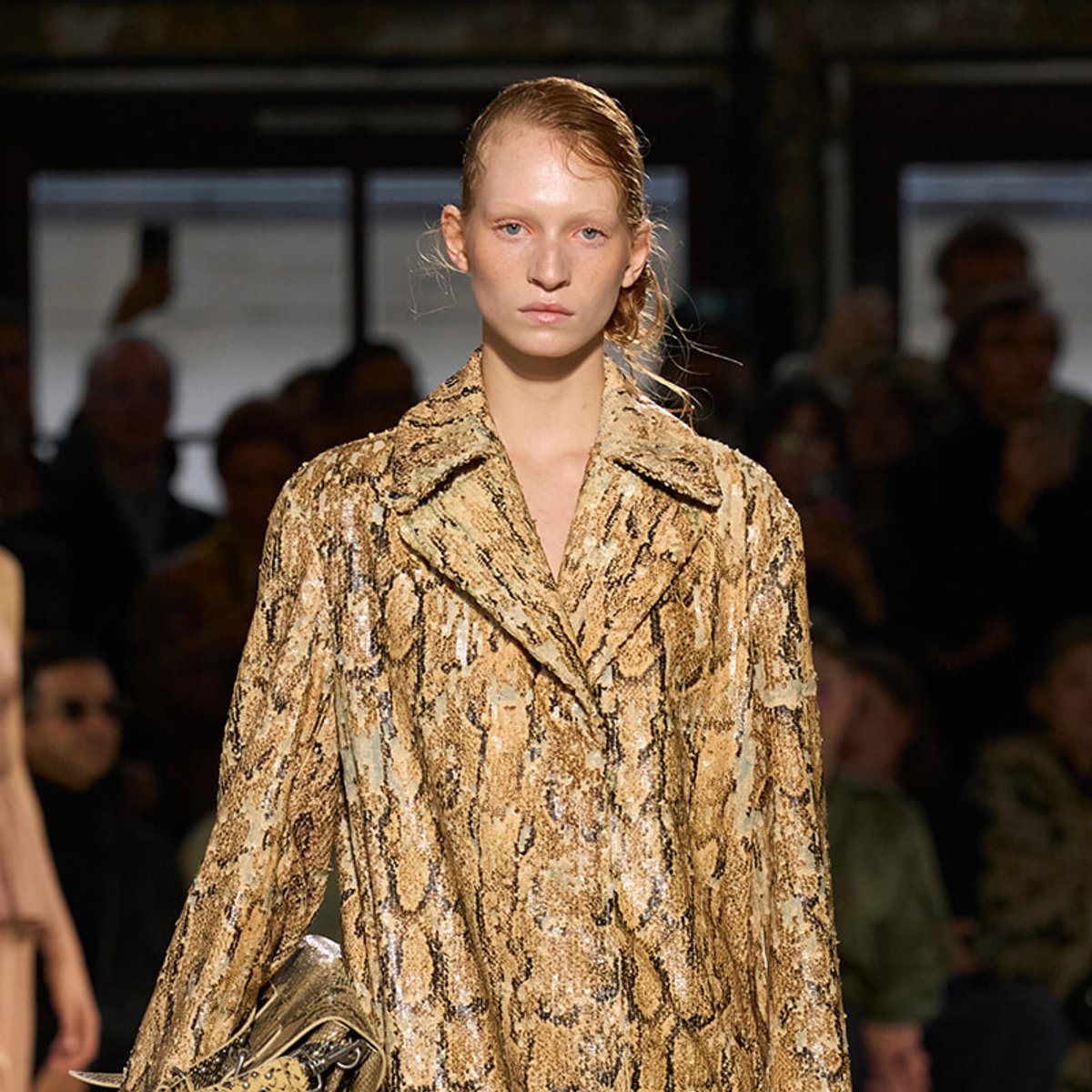 Model walking a fashion runway wearing a gold-brown snakeskin-pattern coat, with a blurred audience in the background.