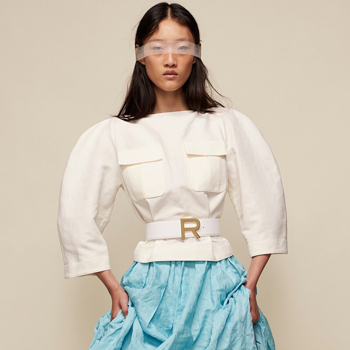 Female model wearing a white top with oversized sleeves, front pockets, and a white belt with a gold R, paired with a light blue voluminous skirt, blindfold across the eyes.