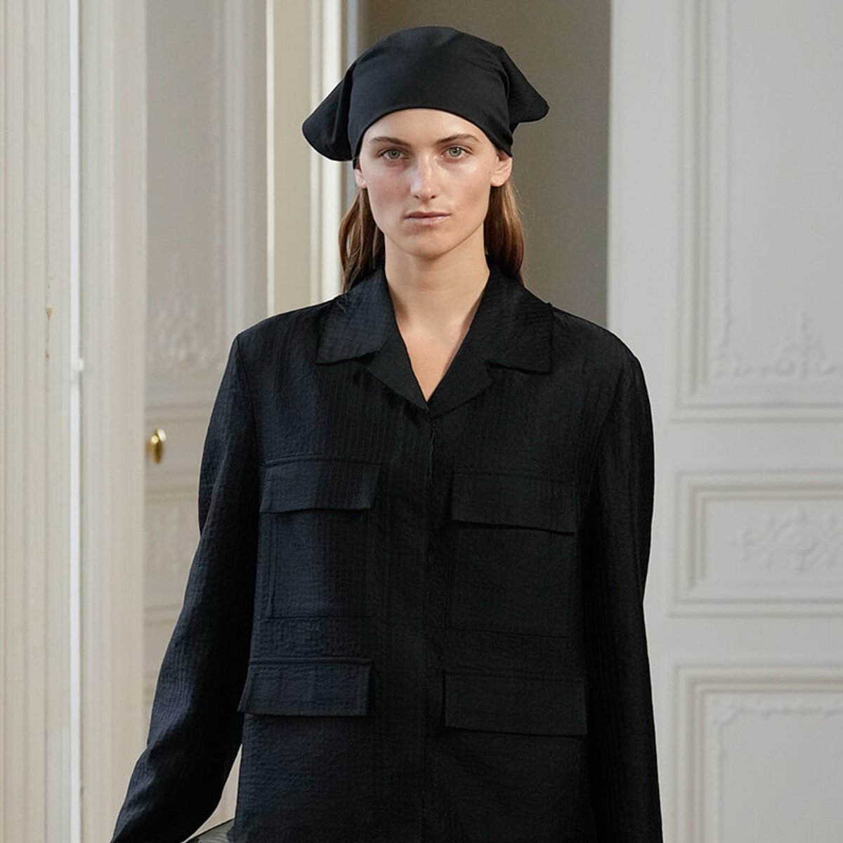 Woman in a black jacket and headwrap stands in a doorway of an elegant room.