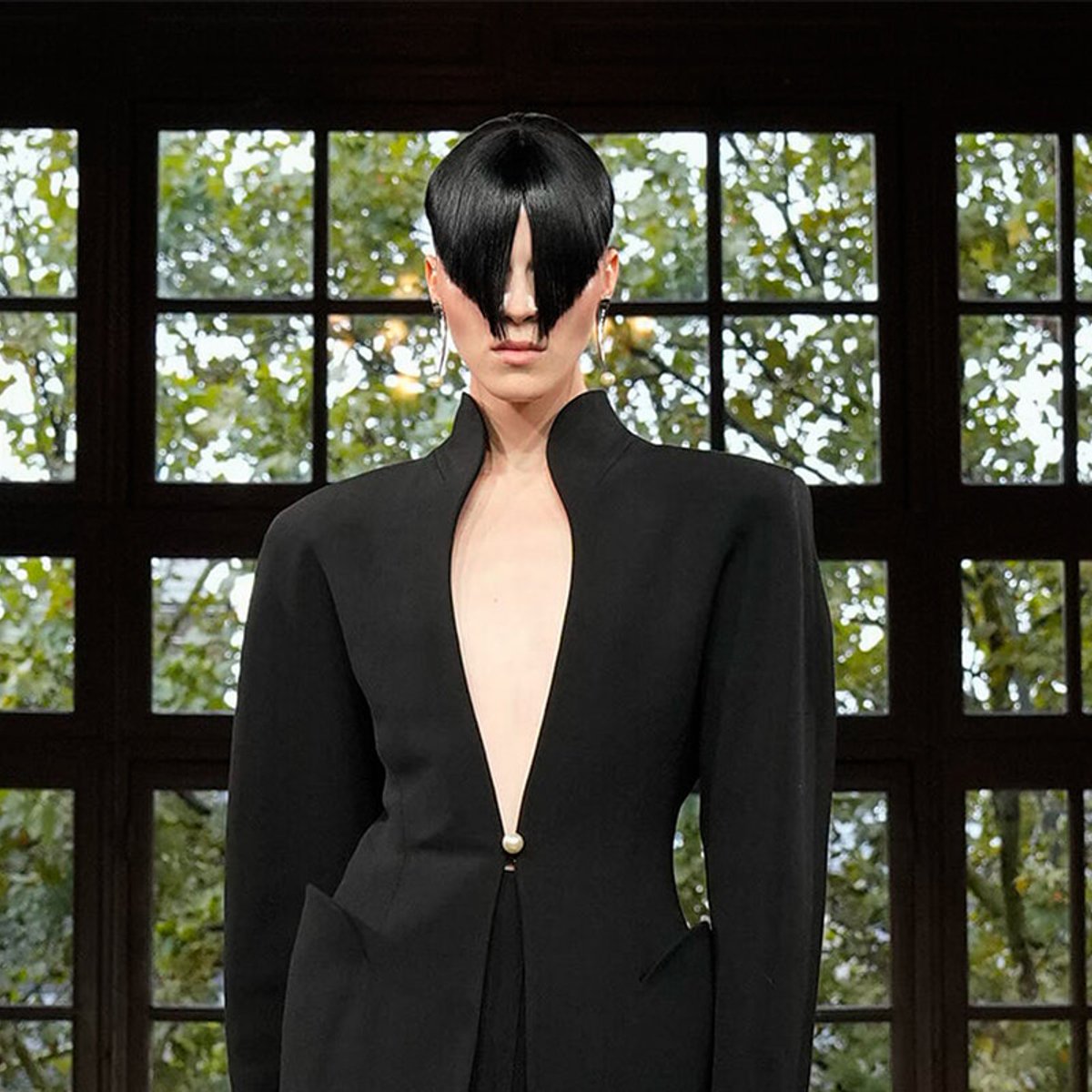 Model on a runway wearing a black tailored outfit with a plunging front and hair swept over eyes, indoors by large windows.