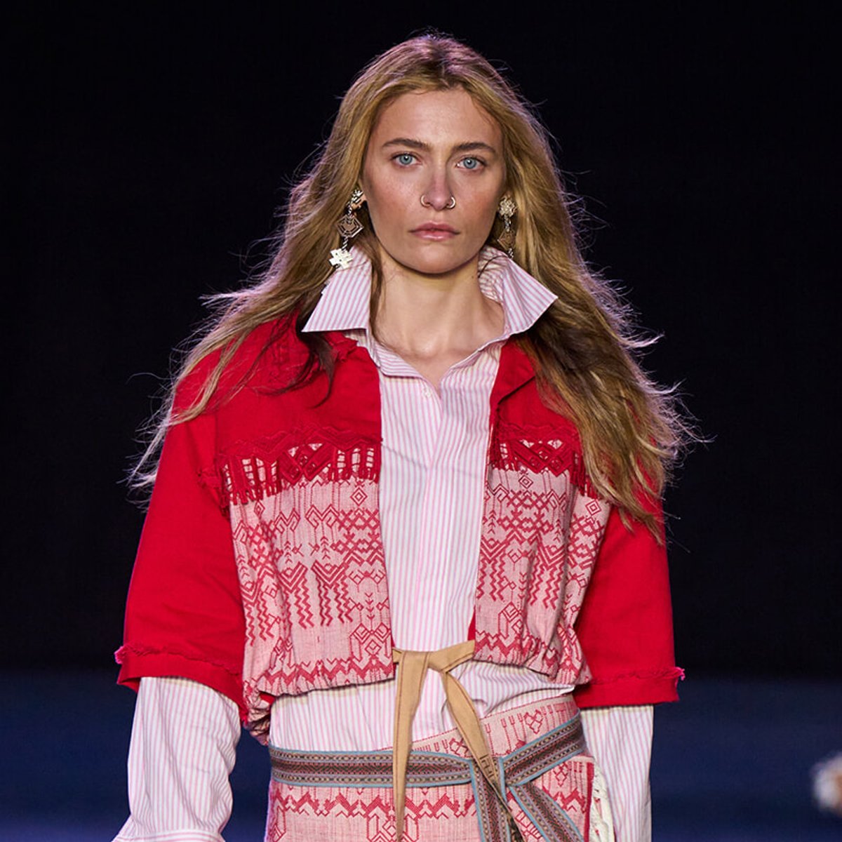Female model on a runway wearing a red patterned jacket over a pink striped shirt with a belt. Has long blonde hair and statement earrings.