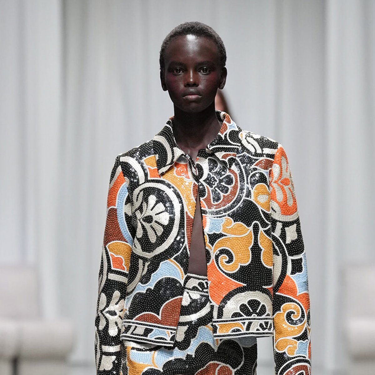 Model wearing a bold multicolored patterned suit with orange, black, white, and blue swirls on a light backdrop, front view on the runway/s runway-style setting.
