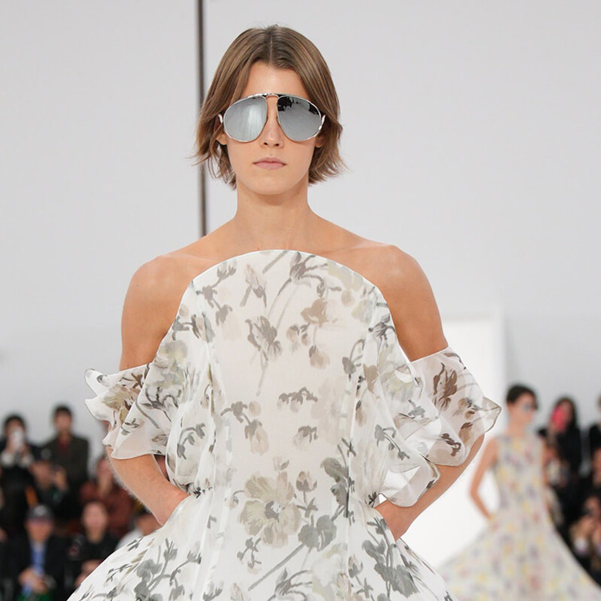 Model in a pale floral off-shoulder dress with ruffled sleeves and oversized mirrored sunglasses on a runway.