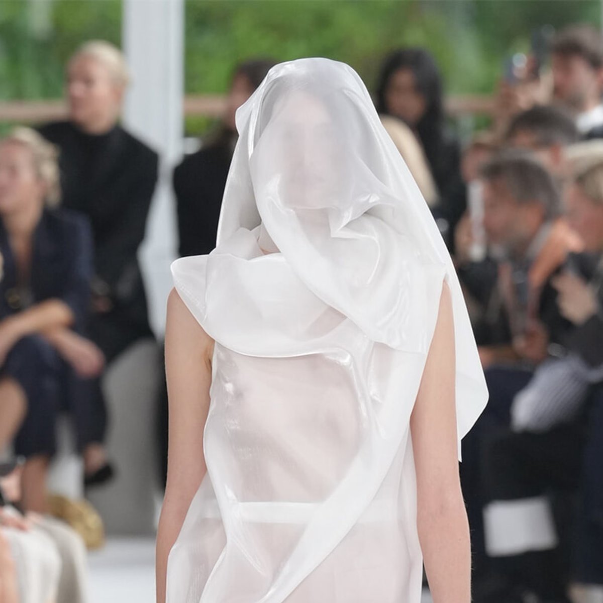 Back view of a model on a runway wearing a flowing white sheer garment with a hood, audience blurred in background.