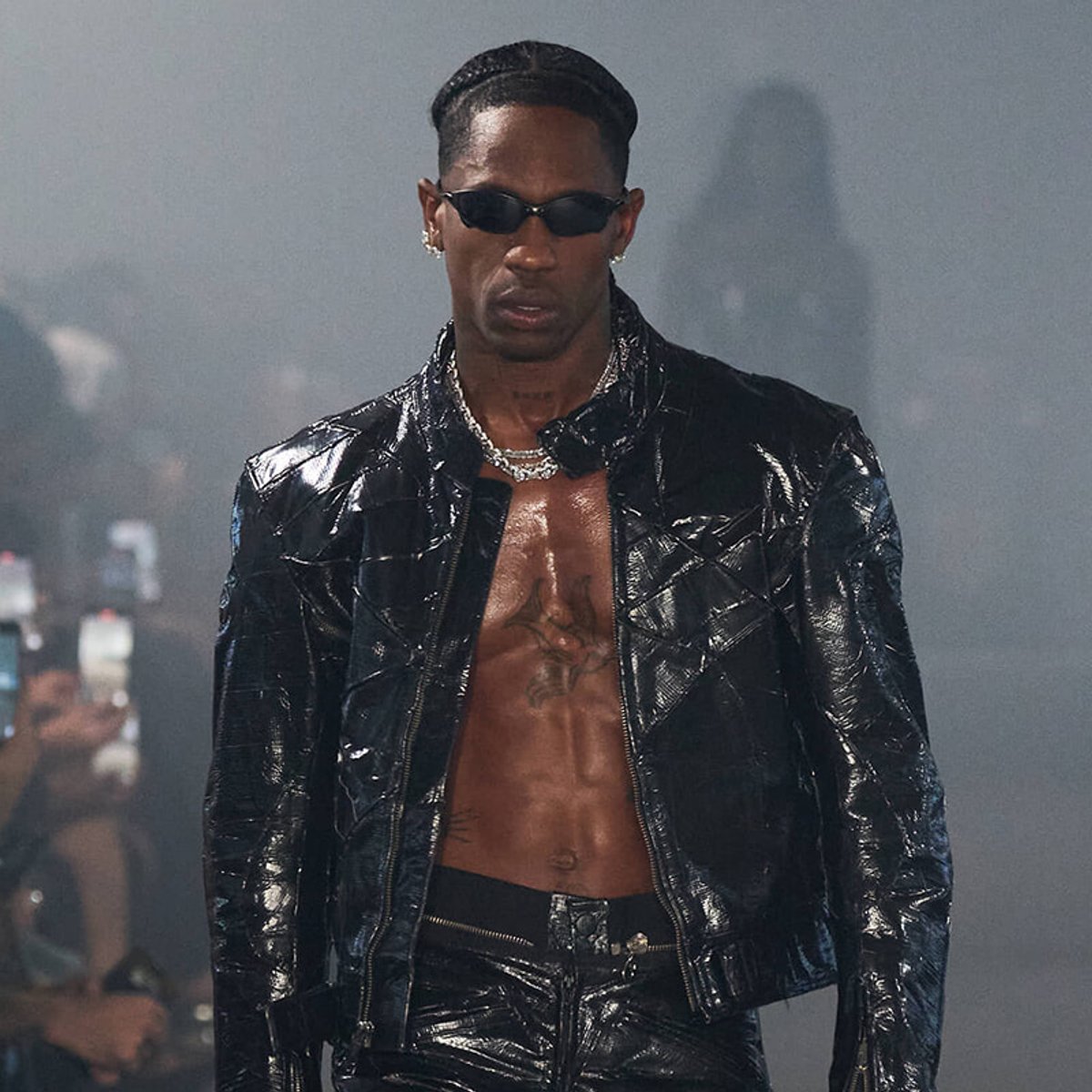 Male model in a glossy black leather outfit with a zipped jacket, sunglasses, and a chain necklace walking on a runway; chest exposed with tattoos visible.