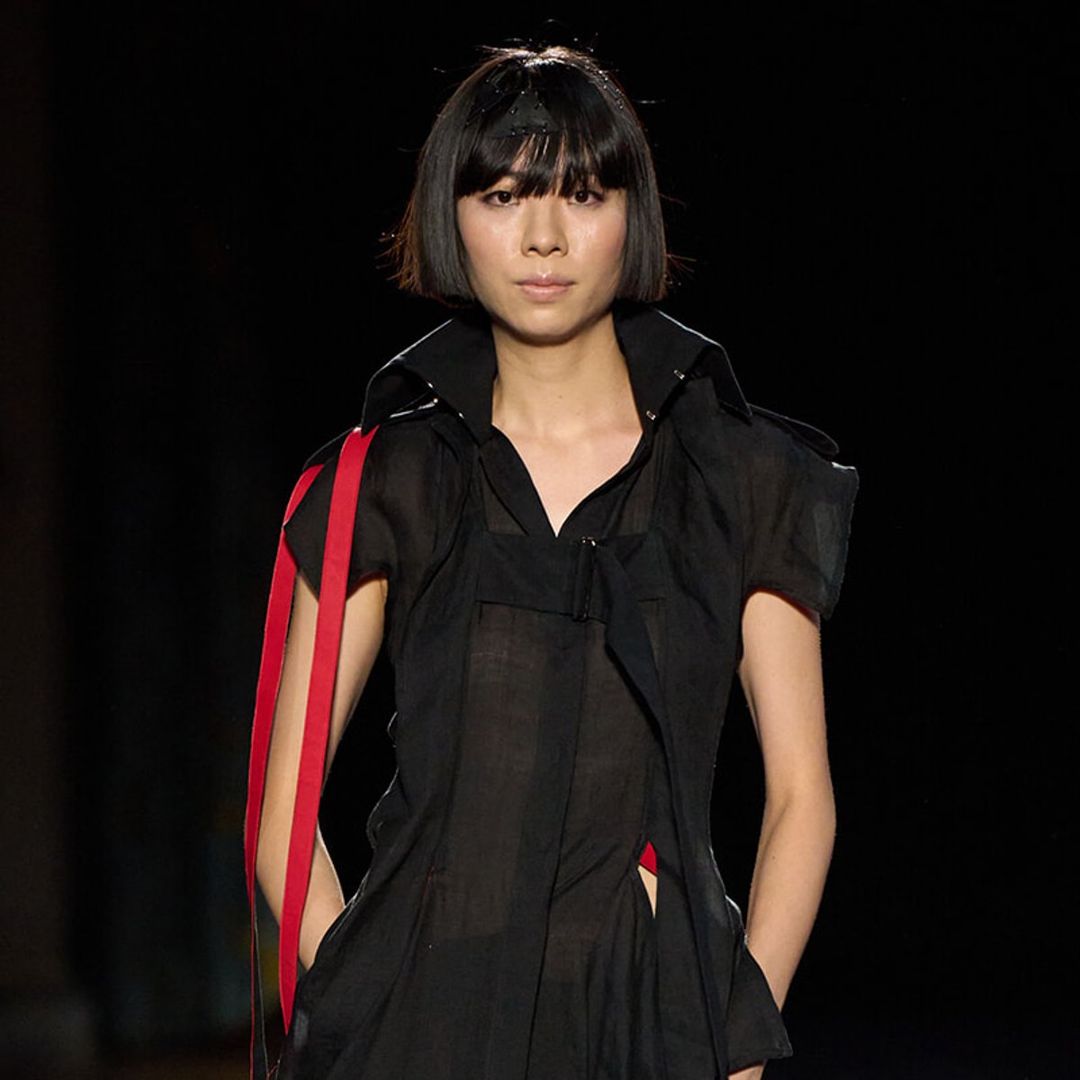 Model walking the runway in a sheer black dress with red shoulder straps.