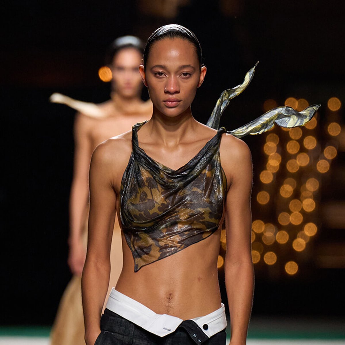 Runway model wearing a camouflage draped crop top with a metallic scarf-like detail over the shoulder, lit catwalk background.