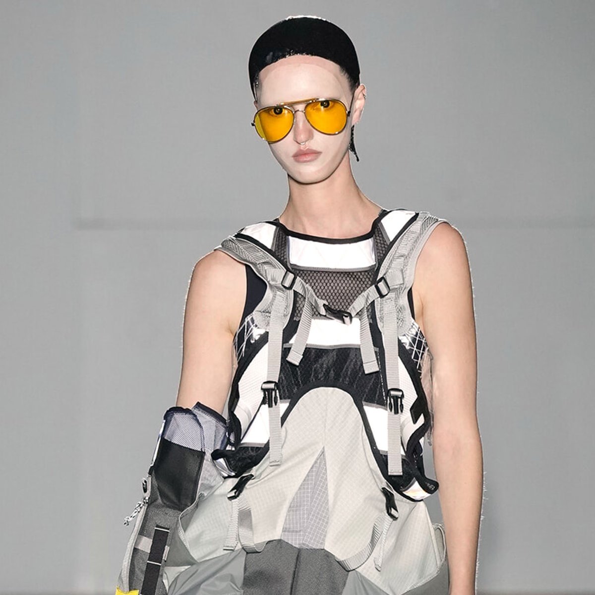 Model wearing a futuristic gray and black harness-style outfit with yellow aviator sunglasses on a runway.