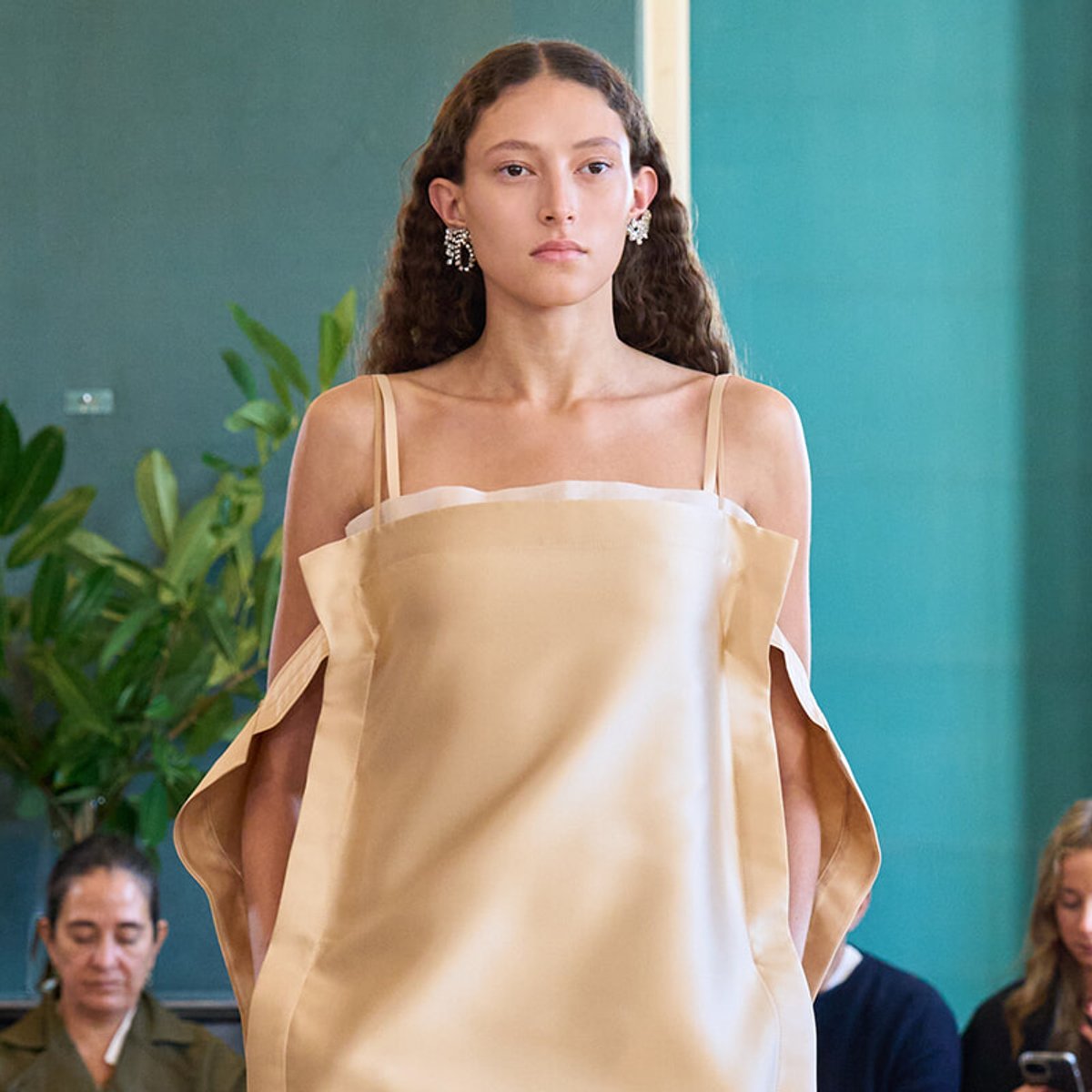 Model walking a runway in a pale satin off-shoulder gown with cape-like sleeves; audience in the background.