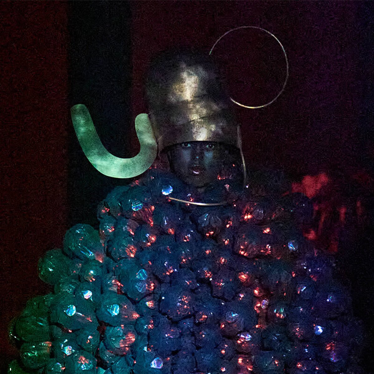 Performer in a metallic helmet with a glowing green horn and a bodice of glowing orb clusters, on a dark stage.
