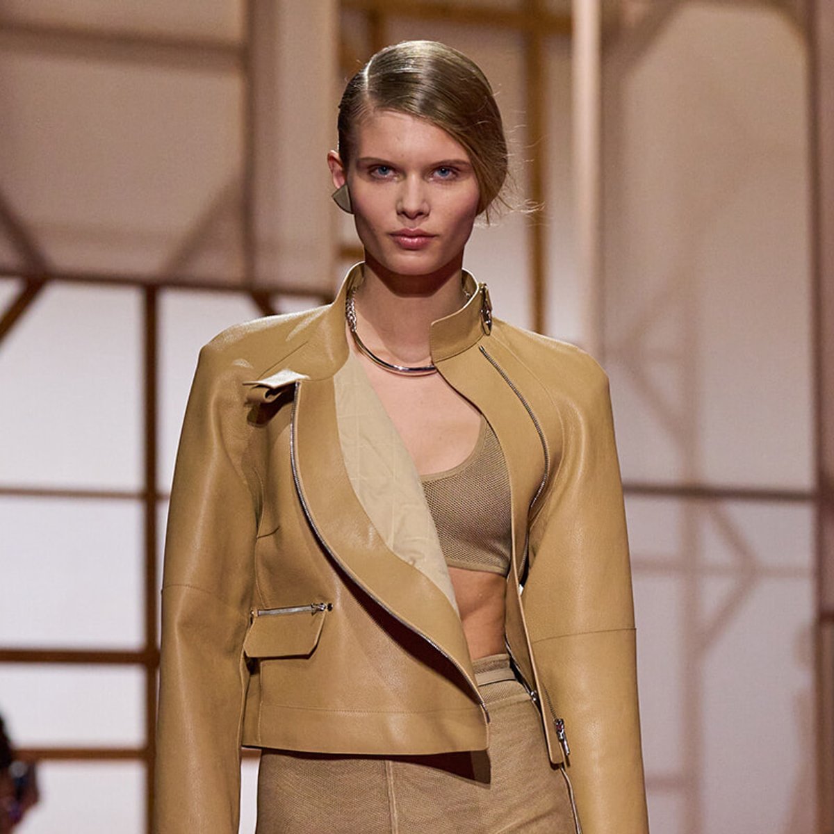 Female model on a runway wearing a tan leather jacket over a beige crop top and matching skirt set, neutral backdrop behind.