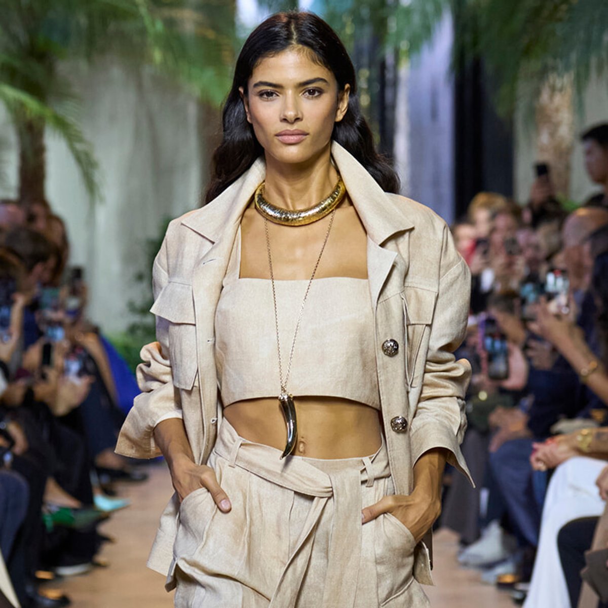 Model walks the runway in a beige two-piece suit with a cropped top and gold chunky necklace.