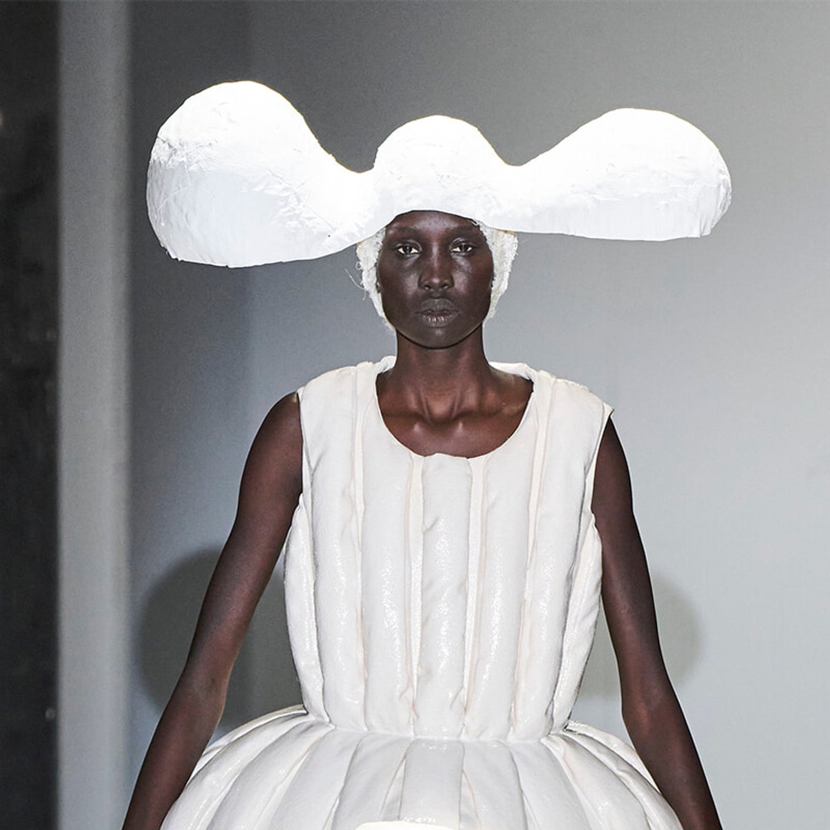 Model walking the runway in an all-white sculptural gown with a large, curved hatpiece resembling a cloud or mushroom cloud.