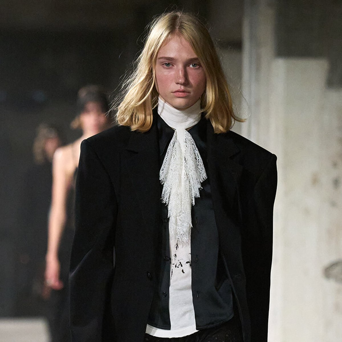 Female model walking a fashion runway in a black blazer with a white lace high-neck scarf.
