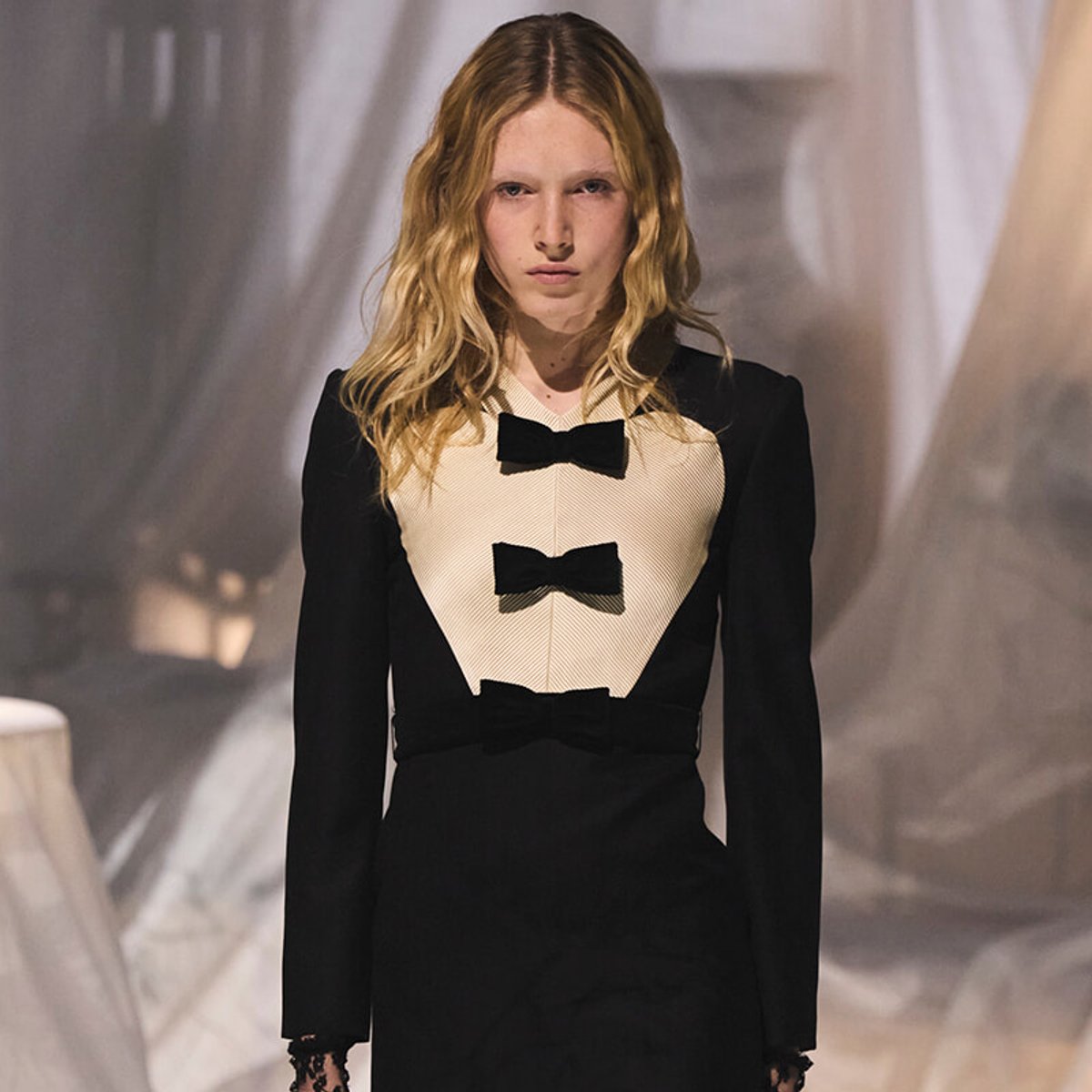 Model walking a runway in a black long-sleeve dress with a cream front panel and three black bow embellishments.