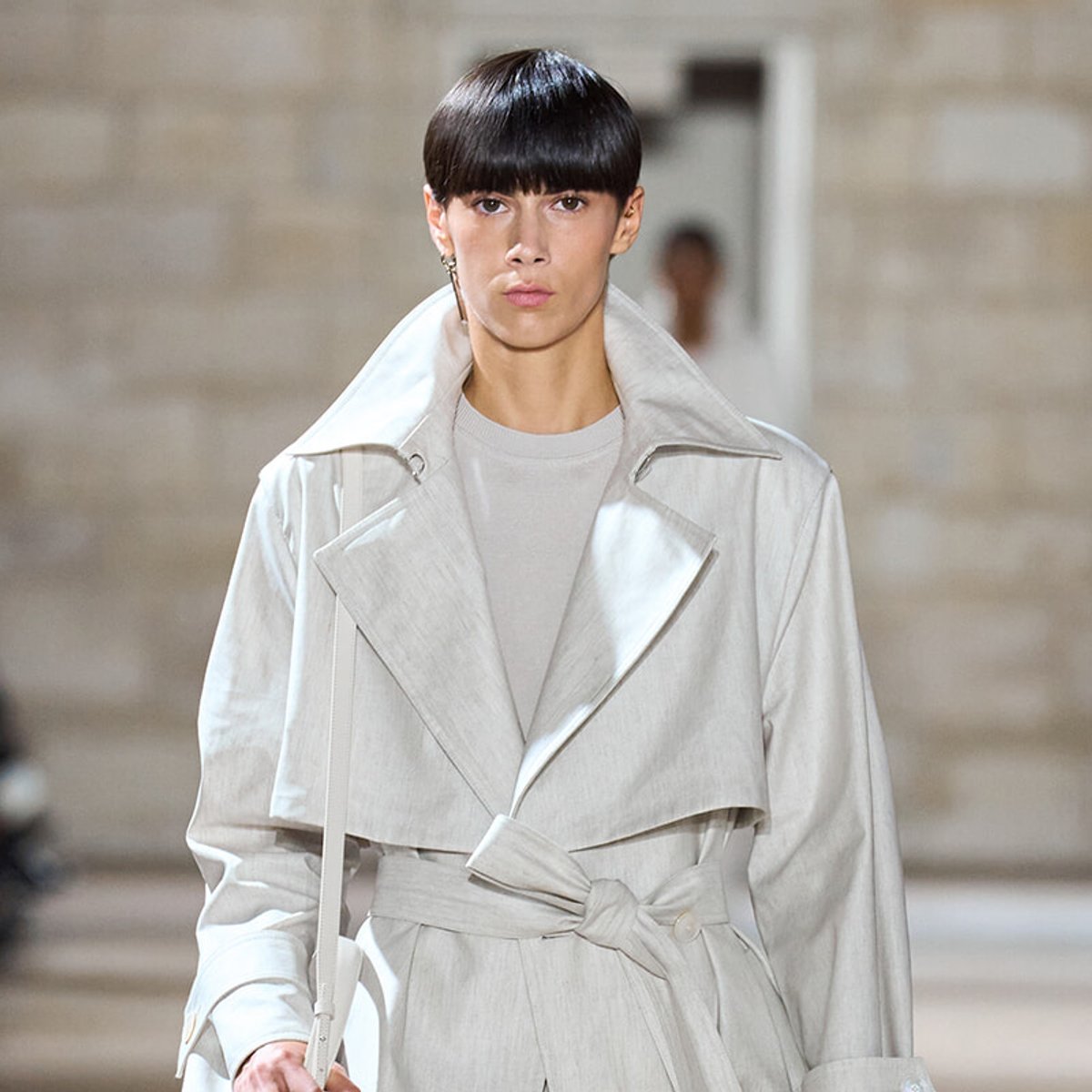 Model walking a runway in a pale gray trench coat with wide lapels and a tied belt at the waist.