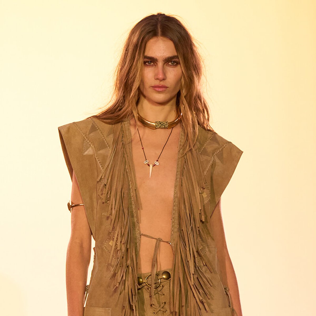 Model on a runway wearing a tan suede fringe vest and boho jewelry against a warm yellow backdrop.