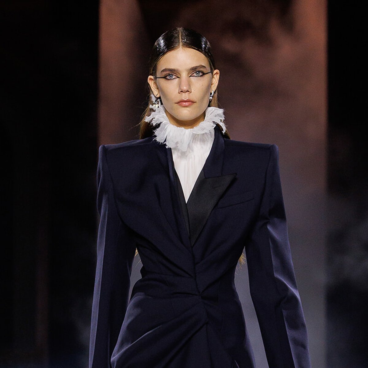 Model walks the runway in a navy tailored blazer with exaggerated shoulders and a white ruffled high-neck blouse, hair slicked back, bold eyeliner.