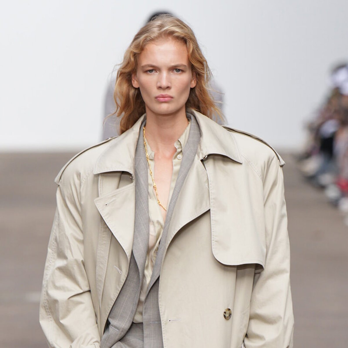 Model walking a runway in a beige trench coat over a pale suit with a gold chain necklace visible.