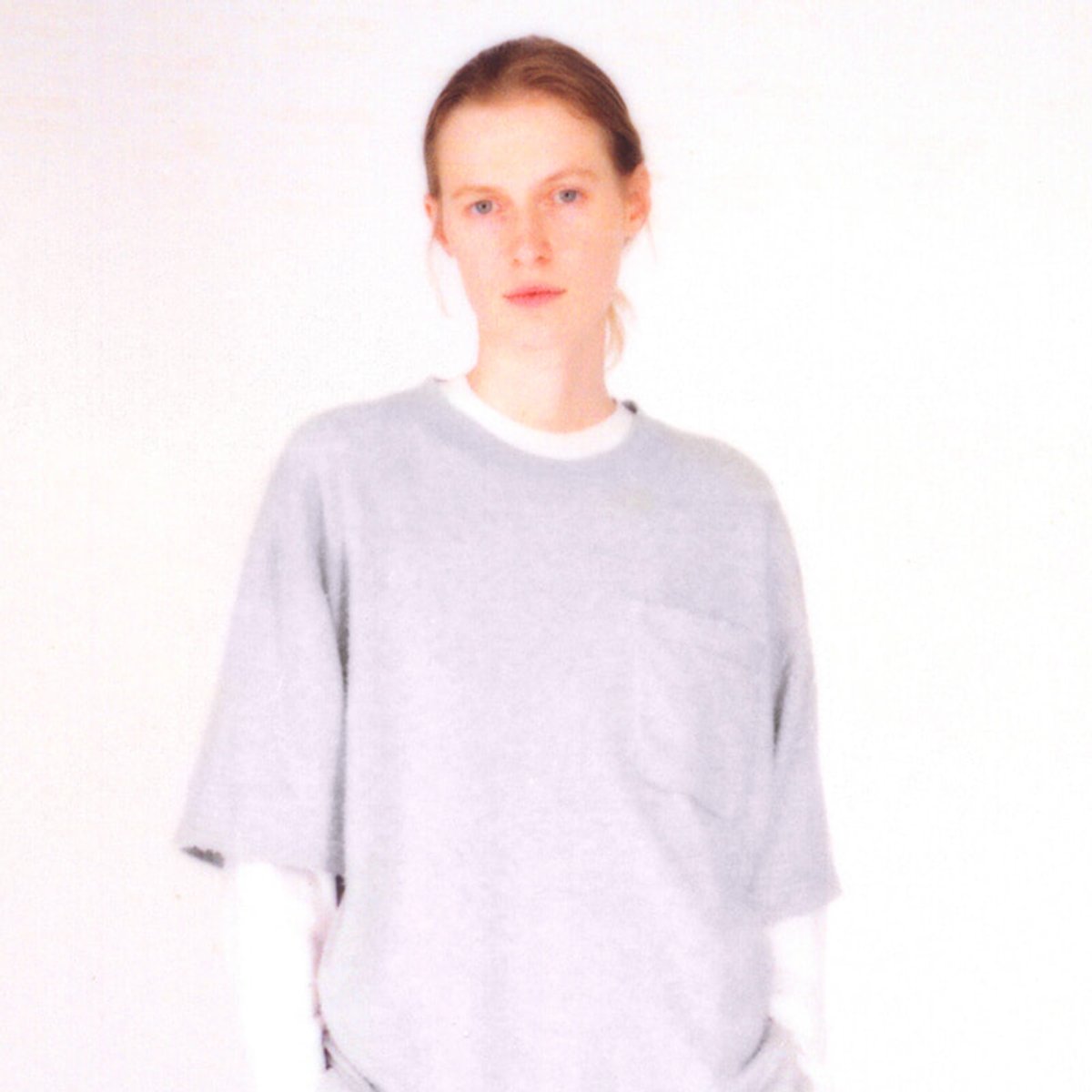 Portrait of a person with light hair wearing a loose gray shirt against a pale background.