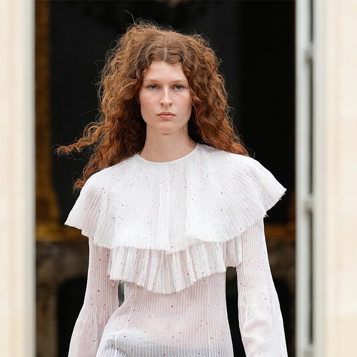 Model with curly red hair wearing a white pleated ruffled blouse standing in a doorway.
