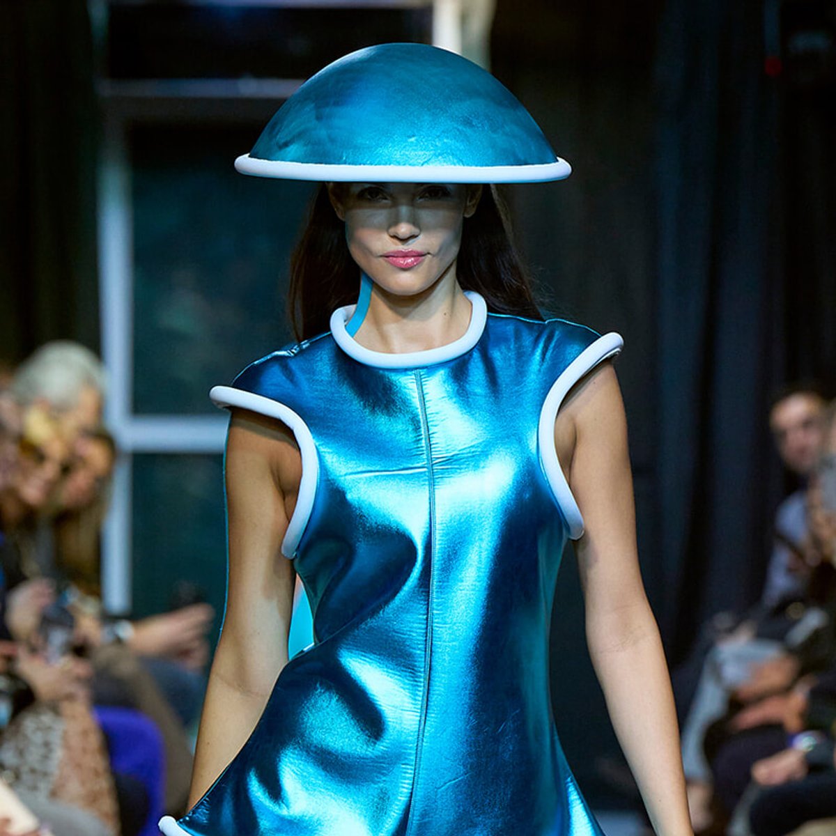 Model walks a fashion runway wearing a metallic blue futuristic dress with white piping and a large blue dome hat.
