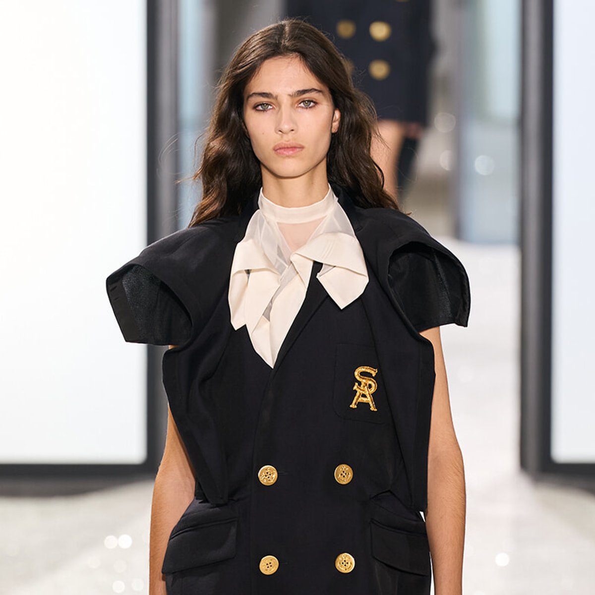 Model walking the runway in a oversized black double-breasted blazer with gold buttons and a cream bow neck scarf.