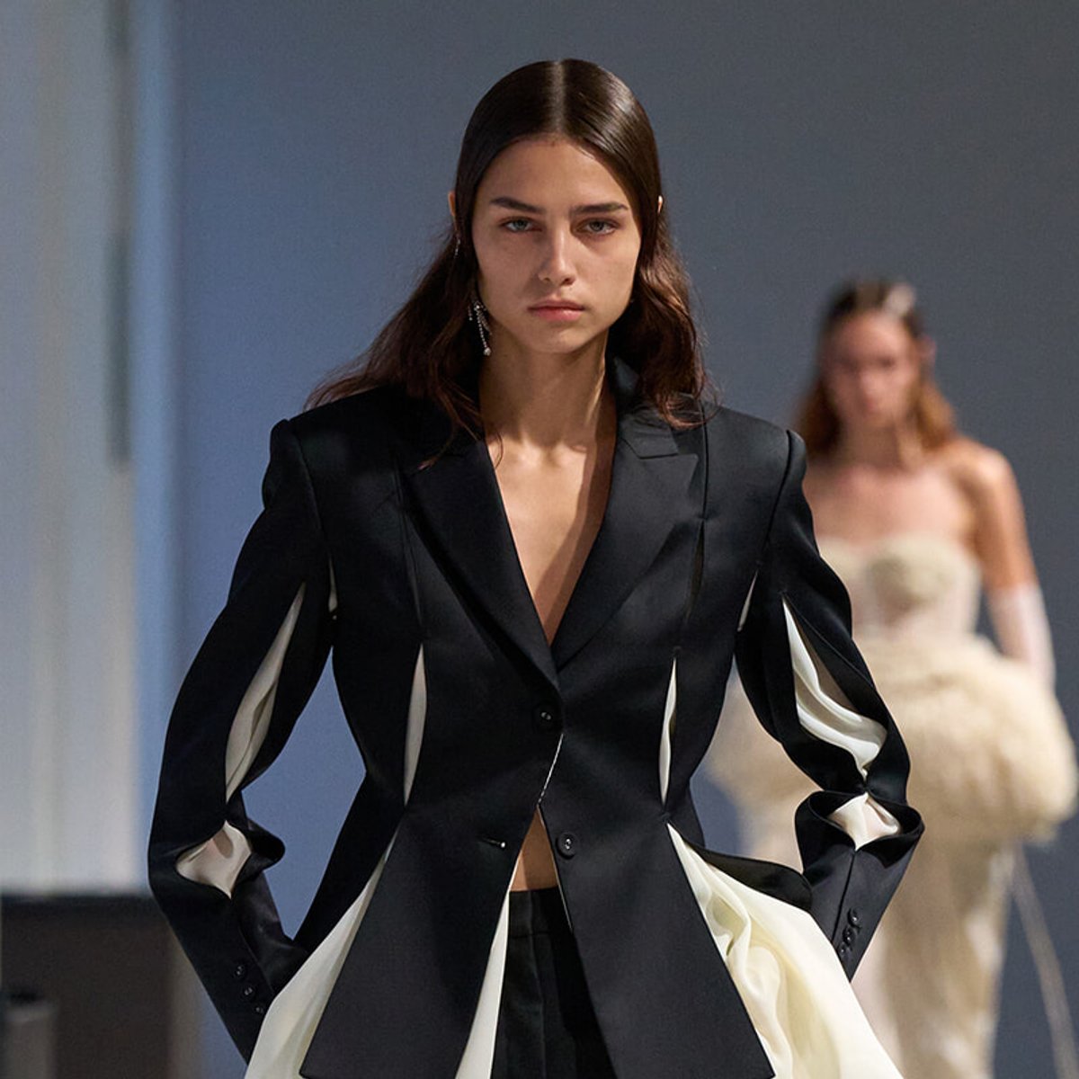 Fashion runway model in a black blazer with white fabric accents, looking forward; another model blurred in the background.