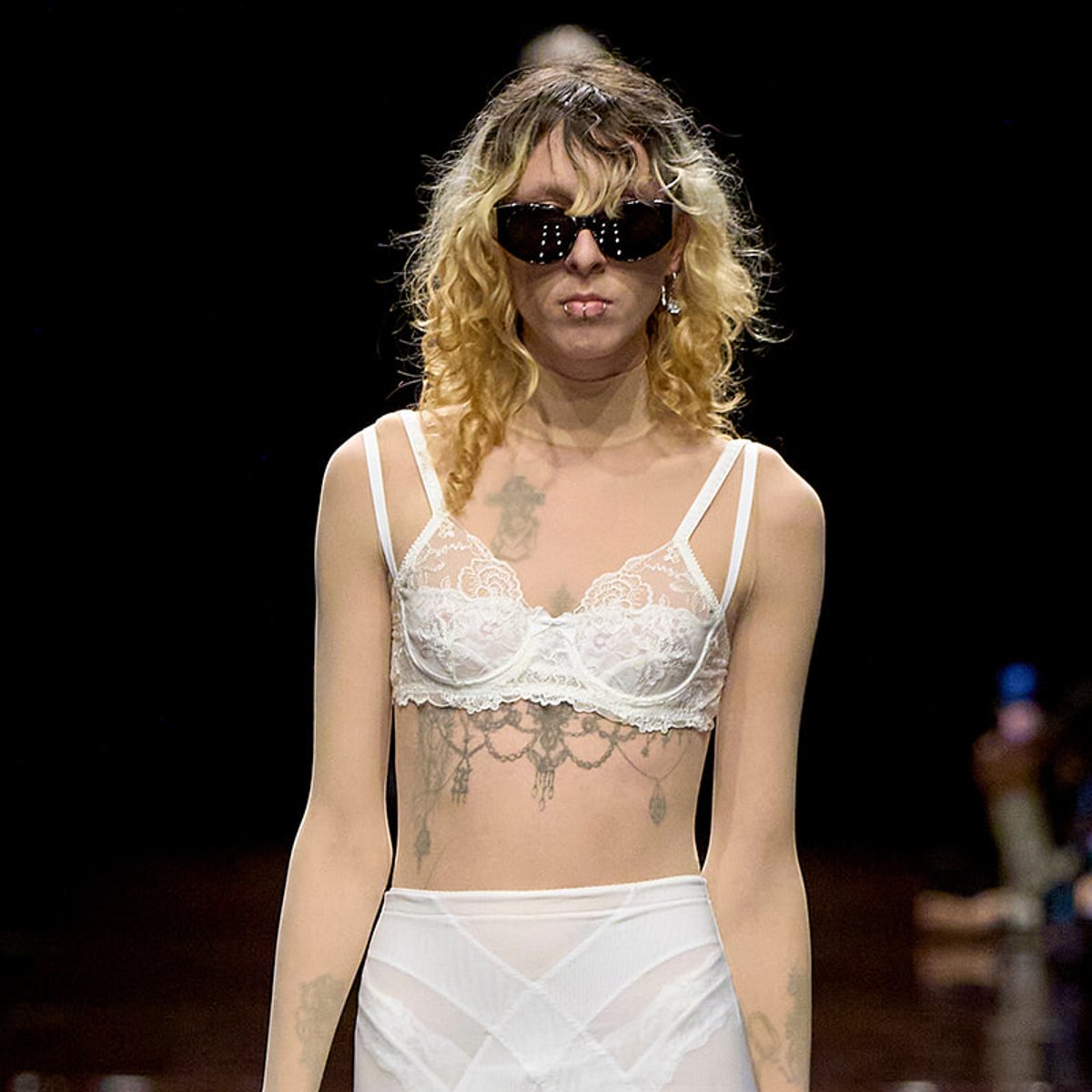 Model walks the runway in a white lace bralette and sheer white underwear, wearing dark sunglasses and visible tattoos.