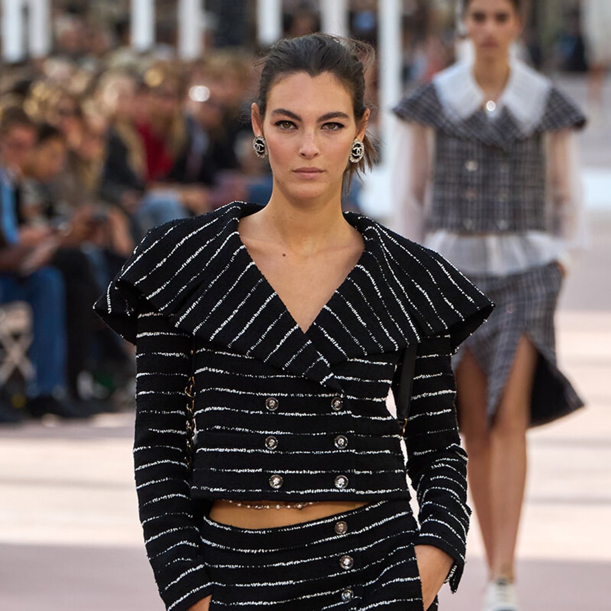 Female model walking a fashion runway in a black and white striped twin-set with structured shoulders, audience blurred in the background.