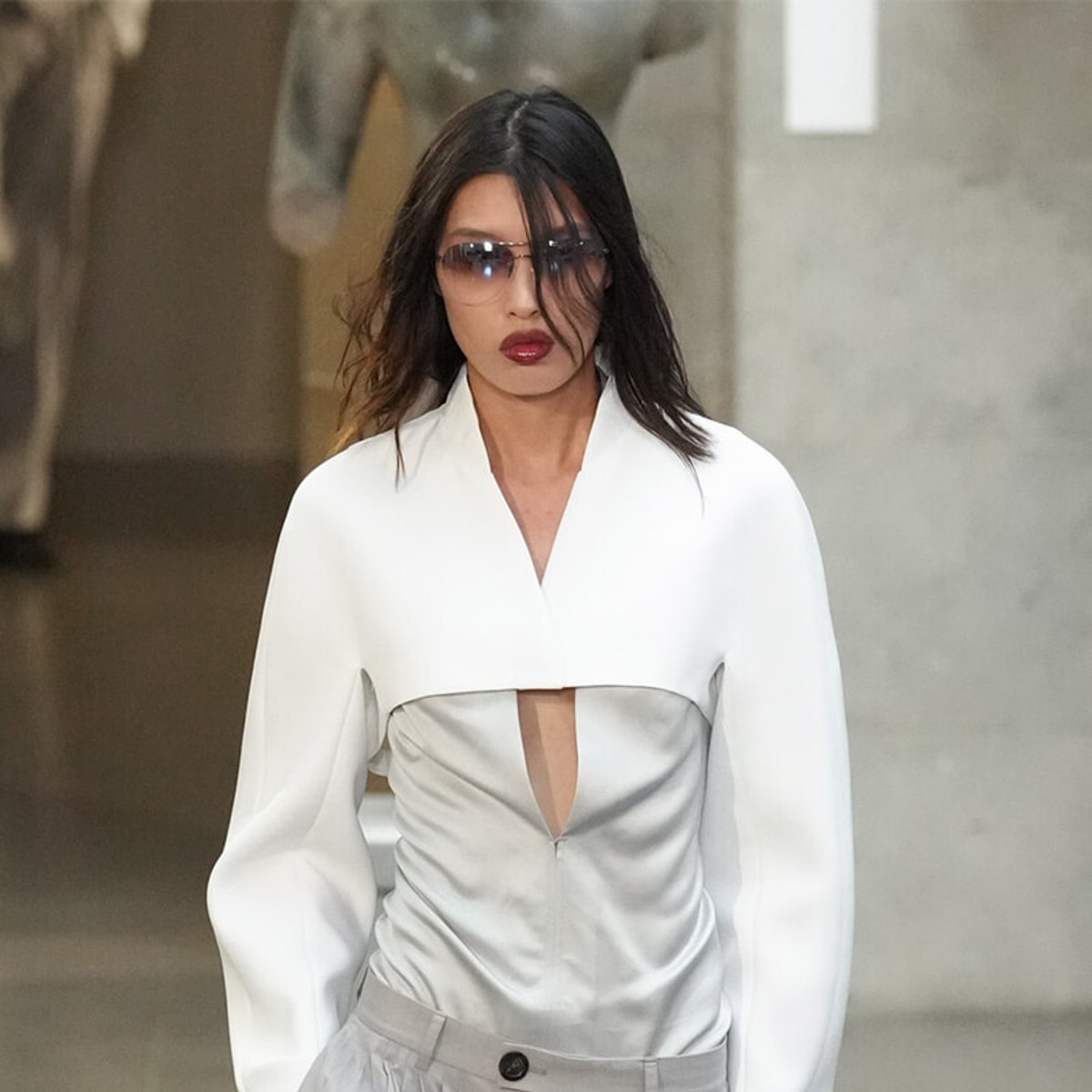 Model walking a runway in a white, cutout-front top and light gray pants, wearing oversized sunglasses.