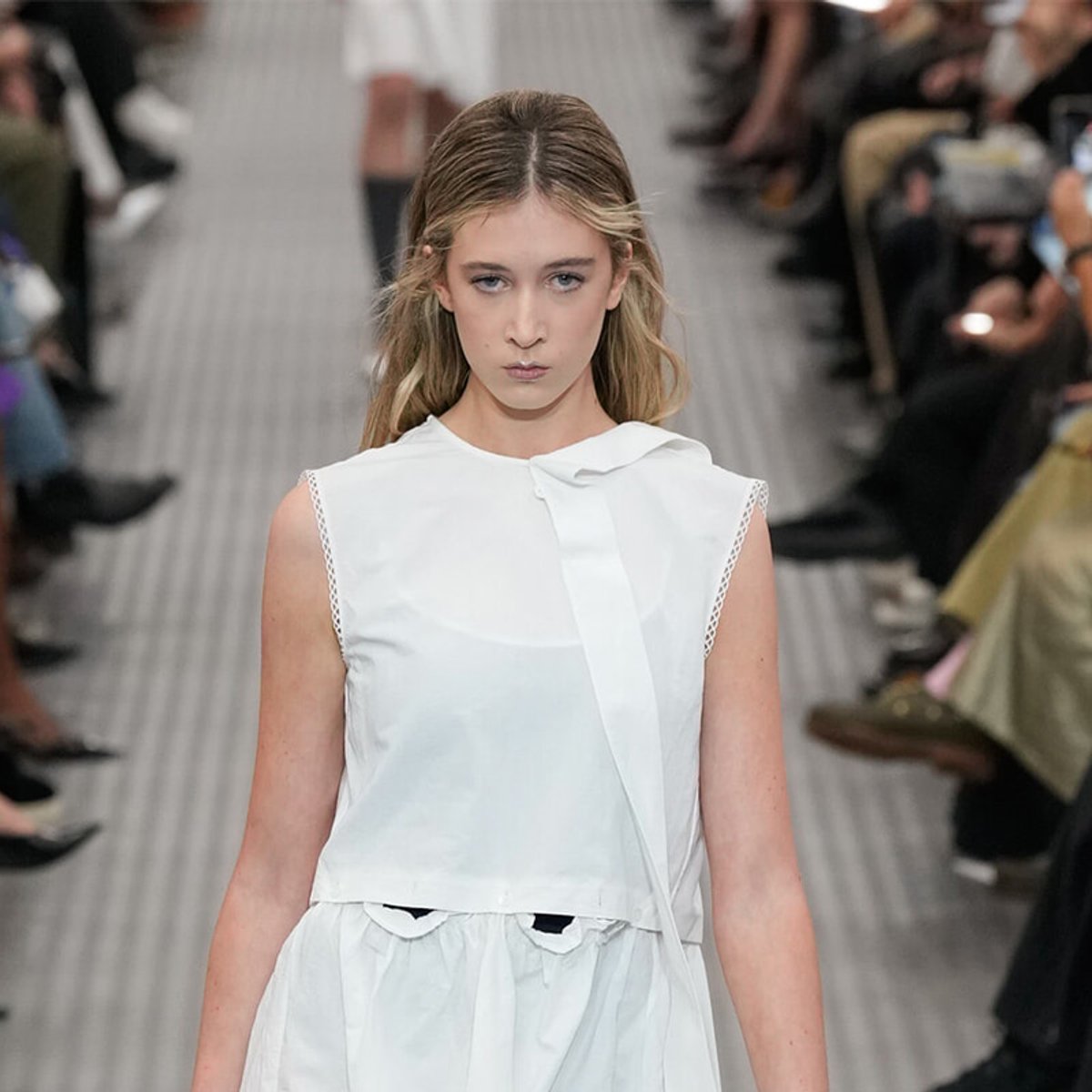 Model walking a fashion runway in a white sleeveless top, focused expression, audience blurred in the background.