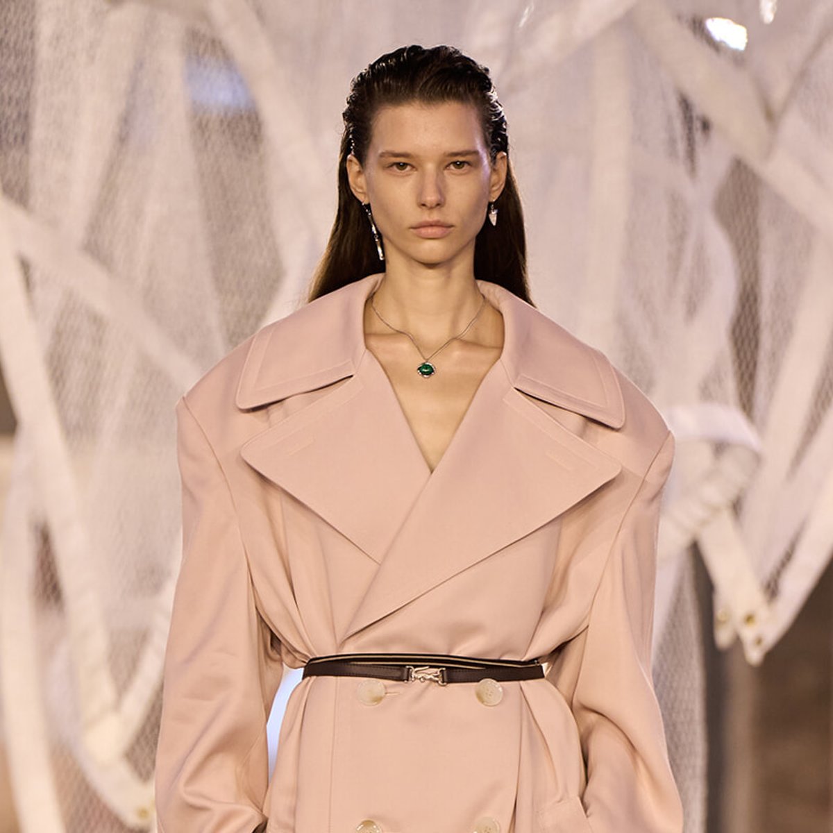 Model on a runway wearing a pale pink trench coat with a black belt and emerald pendant necklace.