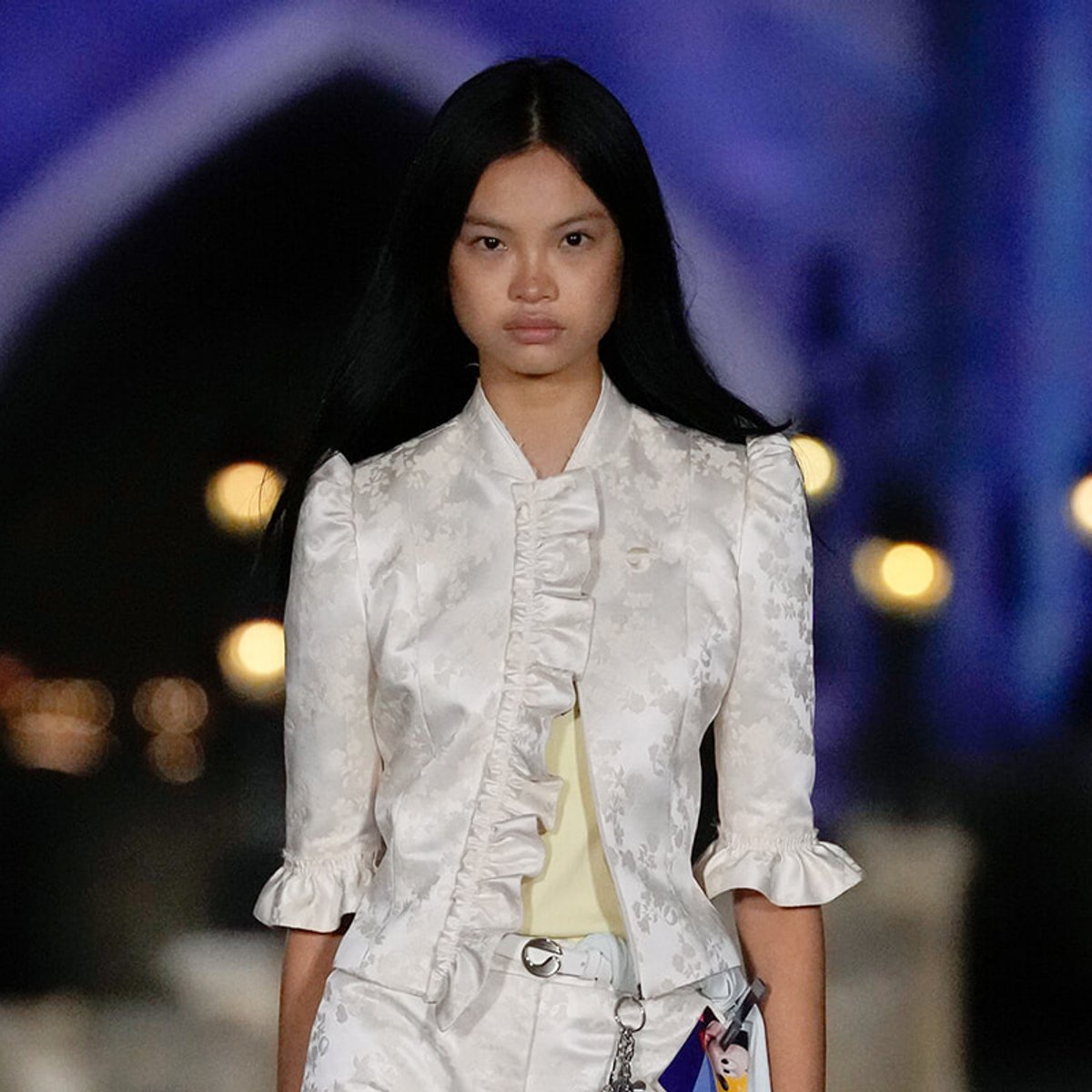 Runway model wearing a white floral blazer with a ruffled front and matching pants, against a blue-lit backdrop.