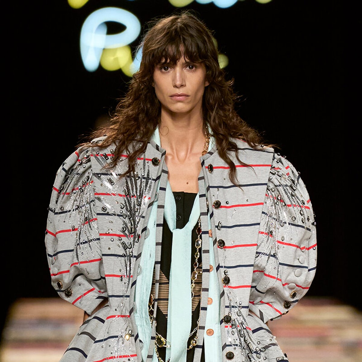 Model on a runway wearing an oversized striped light-gray jacket with red and navy lines, decorated with beads and fringe.