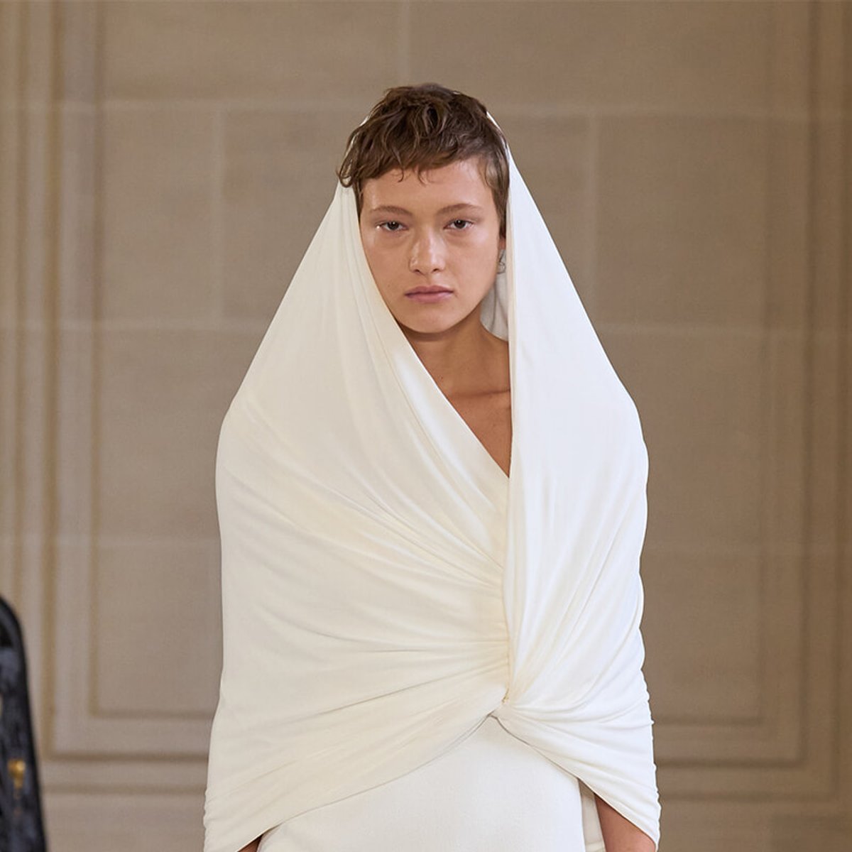 Model on a runway wearing a white draped wrap dress, with fabric wrapped over the head and shoulders, serious expression.