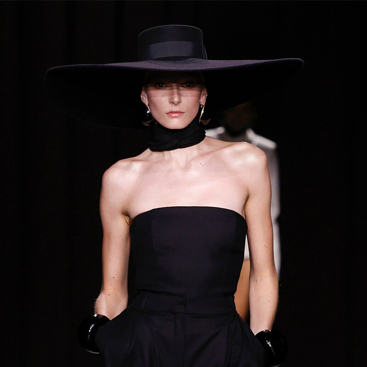 Model on a runway in an oversized black hat and strapless black gown, minimal lighting behind.