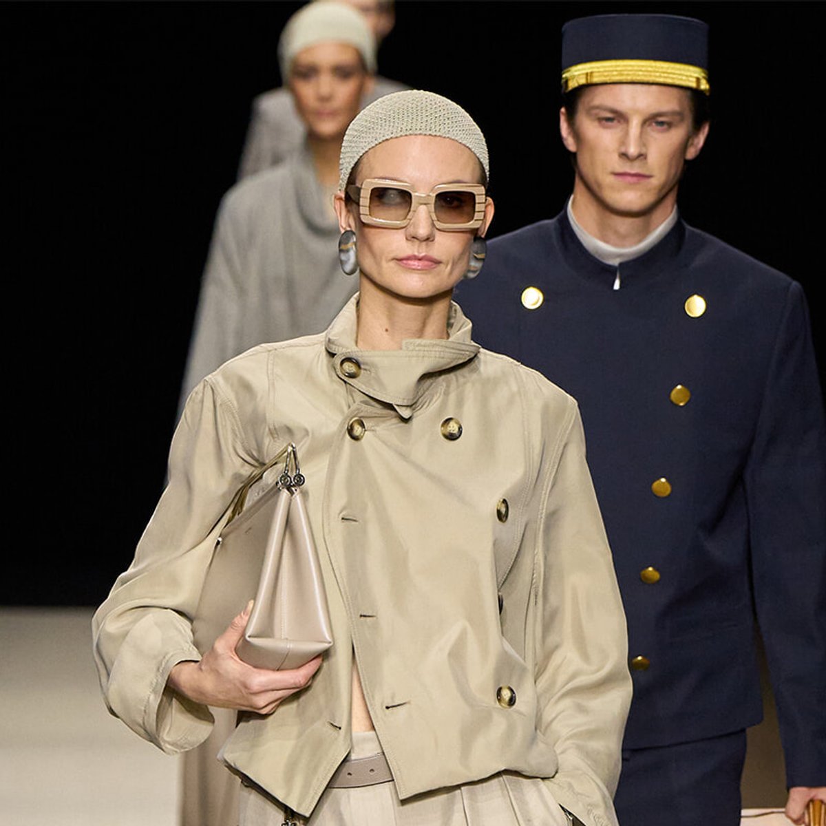 Runway scene with a female model in a beige trench, oversized square sunglasses, and a pale clutch; a navy-uniformed male model with gold buttons walks behind.
