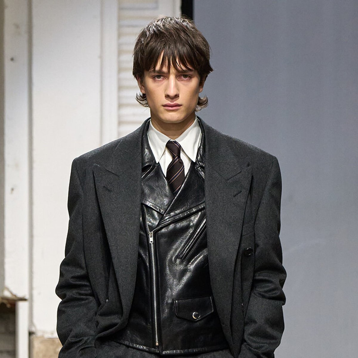 Male model on a runway wearing a charcoal wool coat over a black leather vest, white shirt, and striped tie.