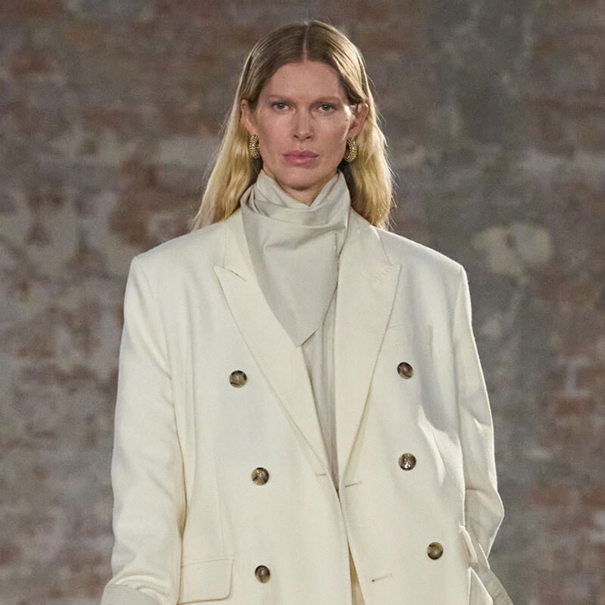 Model walking a runway in a white double-breasted blazer with gold buttons and a light scarf underneath, neutral makeup, brick wall backdrop.