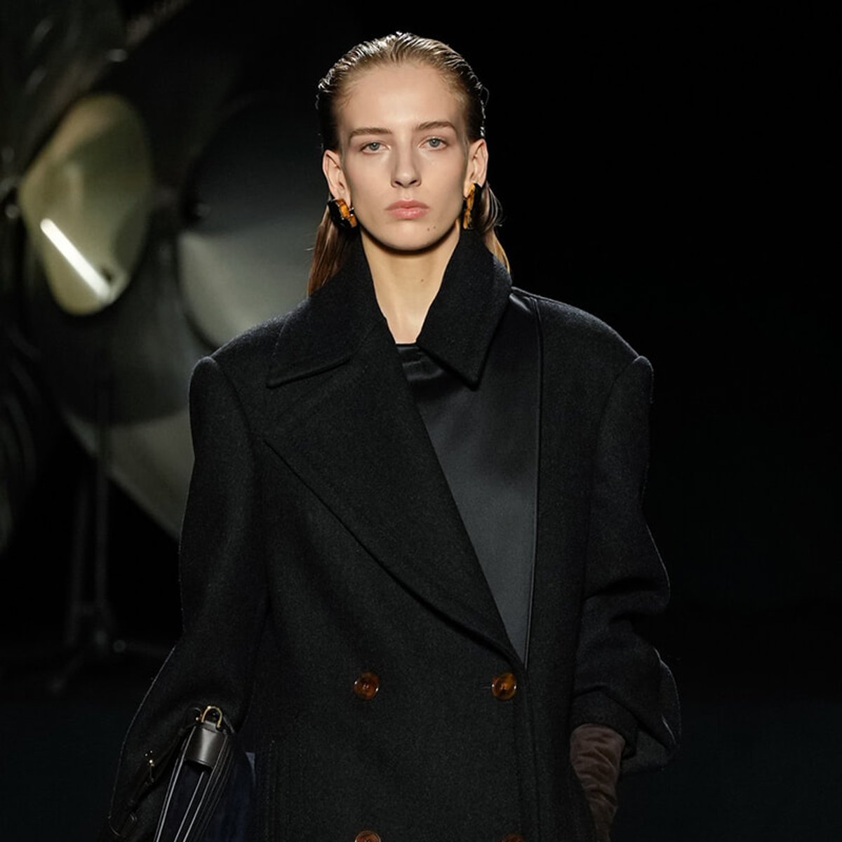 Model walking a runway in a black double-breasted wool coat with large lapels and brown buttons, carrying a bag with a dark strap in her left hand.