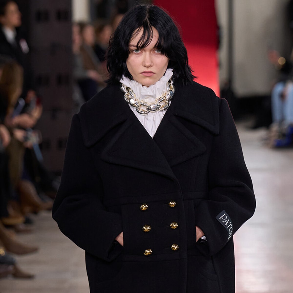Model on a runway wearing a oversized black coat with double gold buttons, a white ruffled blouse, and a chunky pearl necklace.