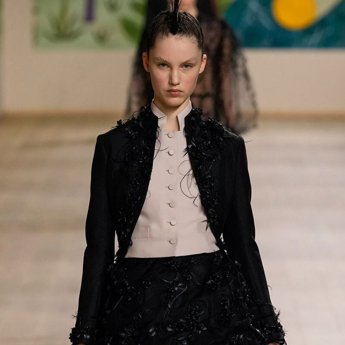 Runway model in a black textured blazer with feathered details over a pale pink buttoned vest, walking the catwalk.