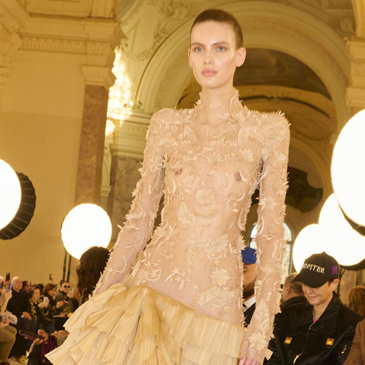 Model walks runway in nude sheer gown with floral appliqué and tiered cream skirt in grand venue with round backstage lights and audience.