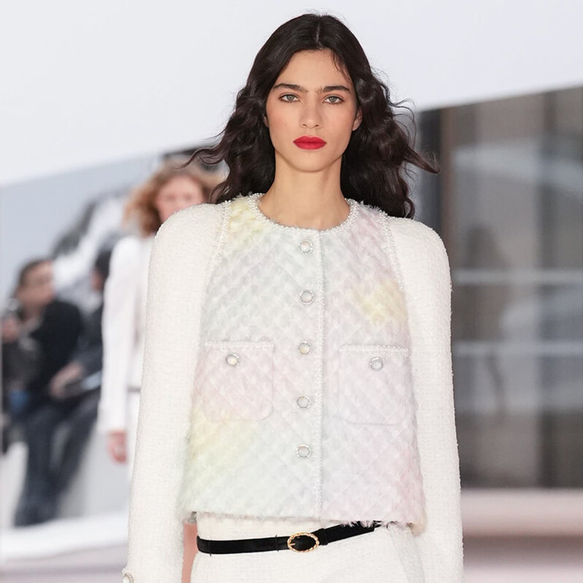 Model walking a runway in a white textured boucle coat with pastel panels, pearl buttons, and a thin black belt.