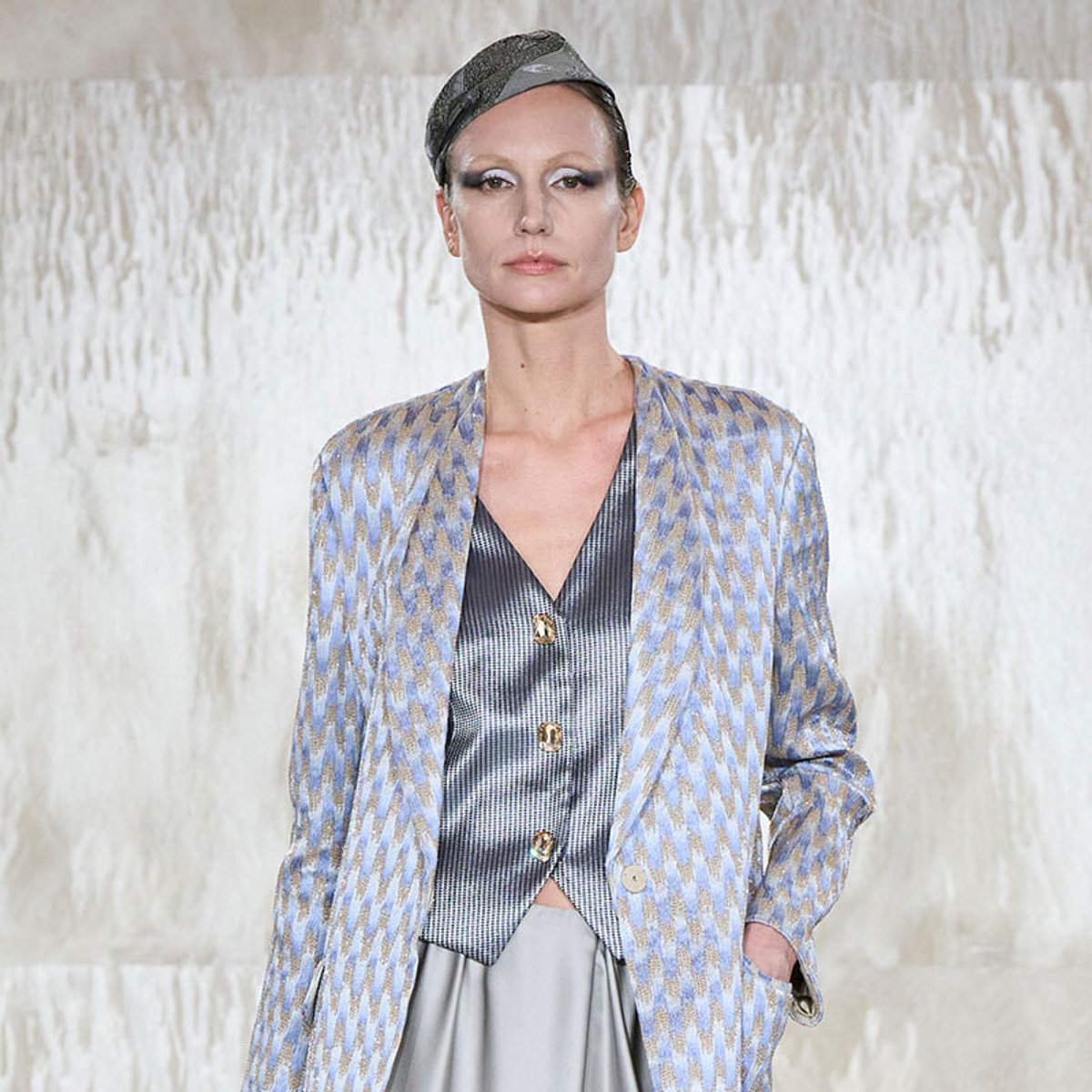 Model on runway wearing a blue zigzag-patterned blazer over a metallic vest and pale skirt, hands in pockets.
