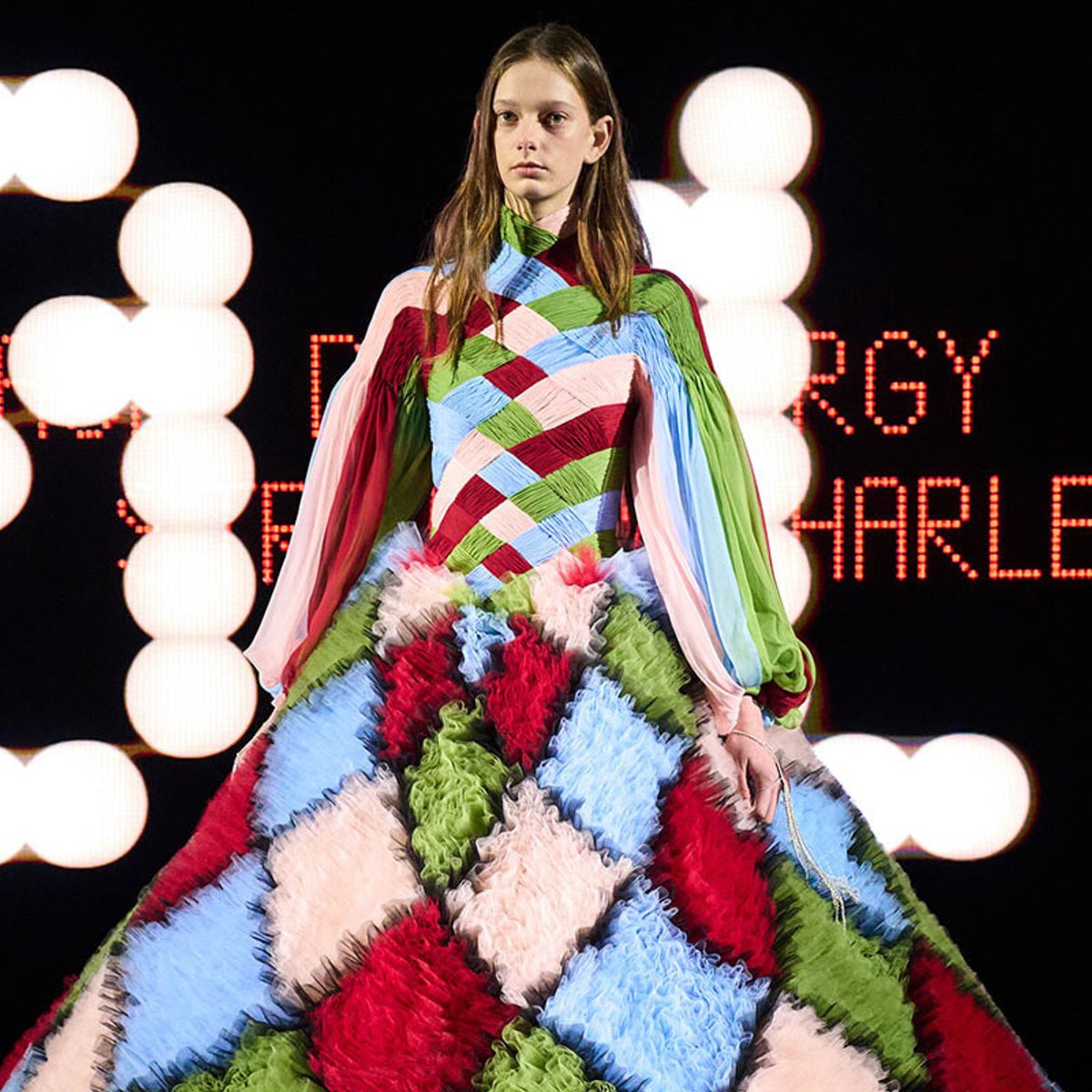 Model walking a runway in a vibrant patchwork gown with crisscross ribbons and a voluminous, ruffled skirt against a backdrop of bright stage lights.