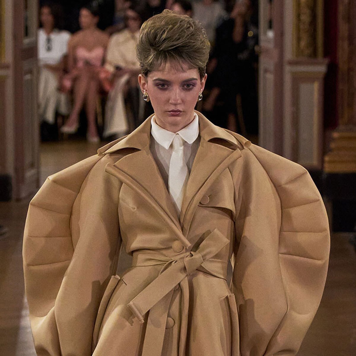 Model on a runway wearing an oversized tan trench coat with dramatic puff sleeves and a white shirt underneath.